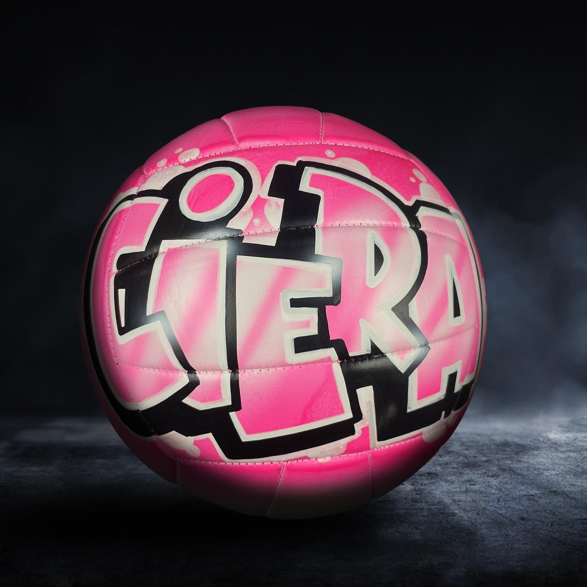 A pink and black volleyball with graffiti-style text on it, sitting on a dark surface.