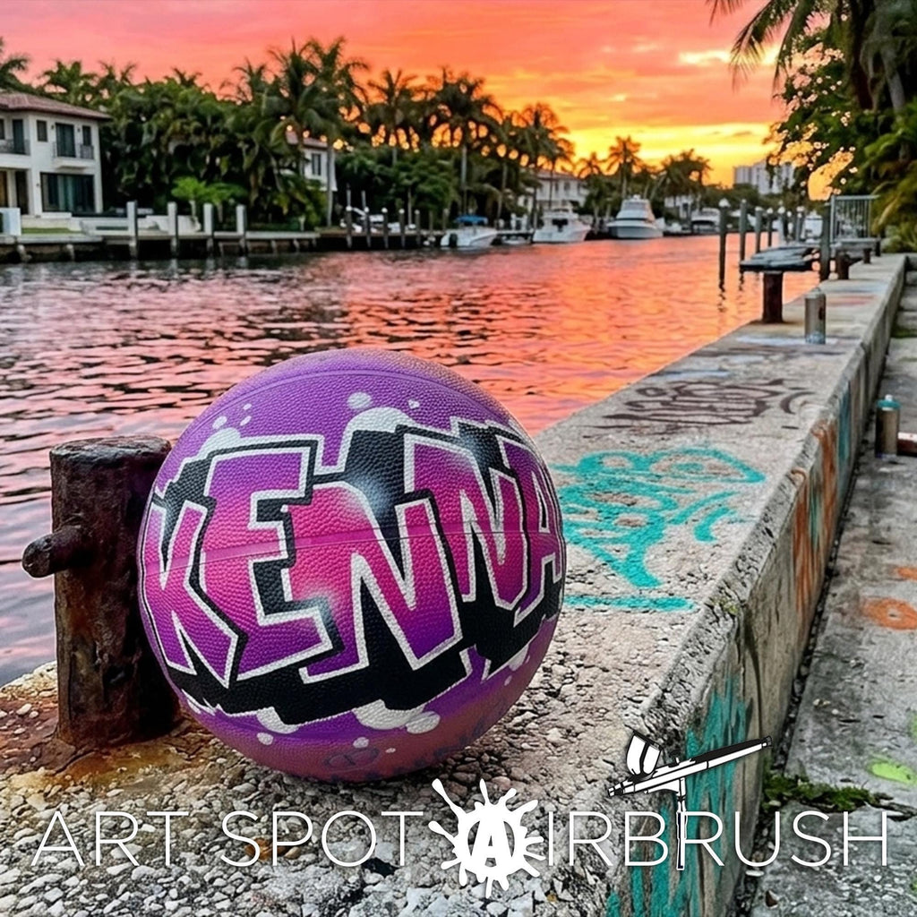 A colorful basketball with the word "KENYA" painted on it, sitting on a concrete ledge near a body of water with palm trees and a sunset in the background.