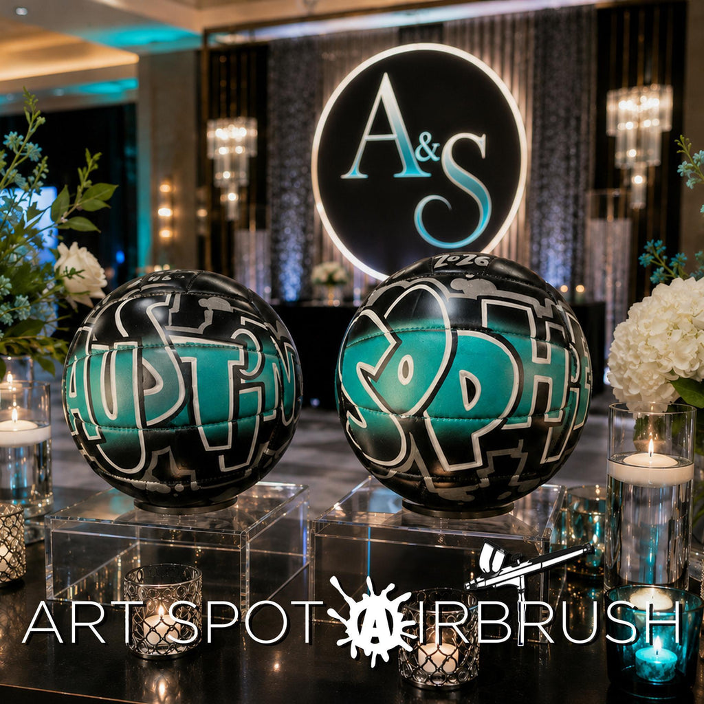 two large, black and teal graffiti-style custom volleyball with the names "SOPHIE" and "AUSTIN" written on them, placed on a table with candles and flowers. In the background, there is a large circular logo with the letters "A&S" and a number "2016". The image also includes the text "ART SPOT AIRBRUSH".