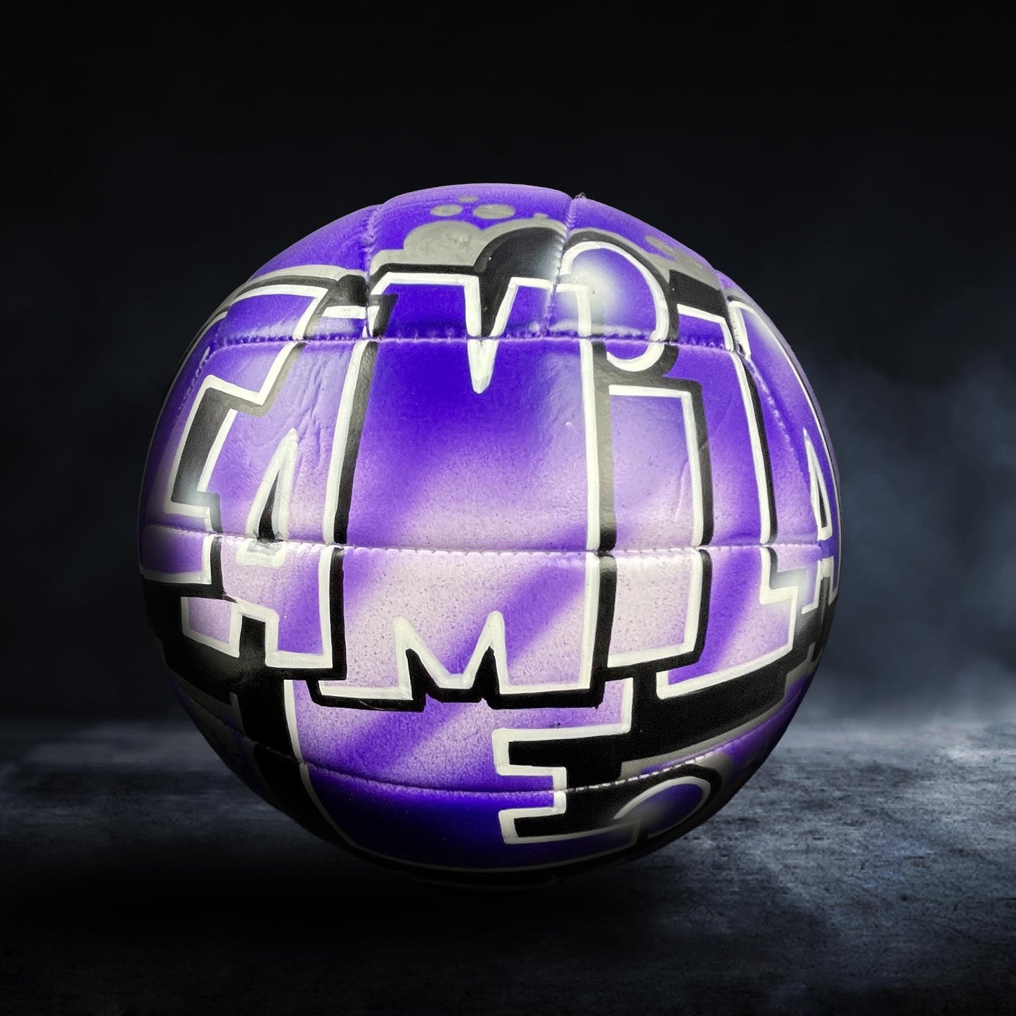 A purple and black soccer ball with graffiti-style text on it, set against a dark background.