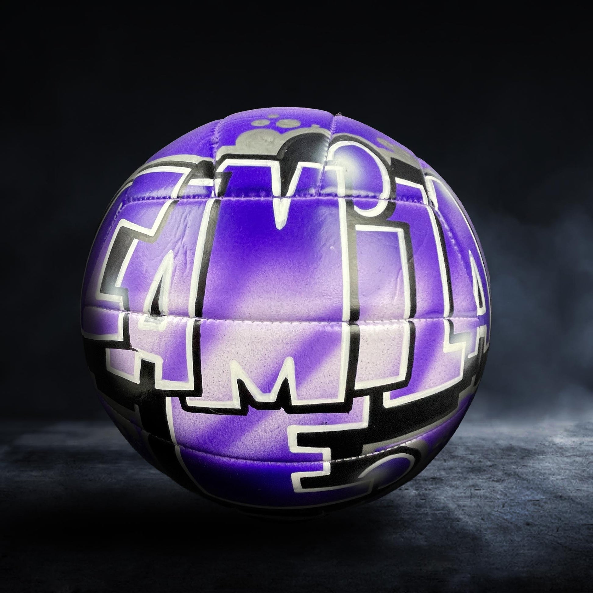 A purple and black soccer ball with graffiti-style text on it, set against a dark background.