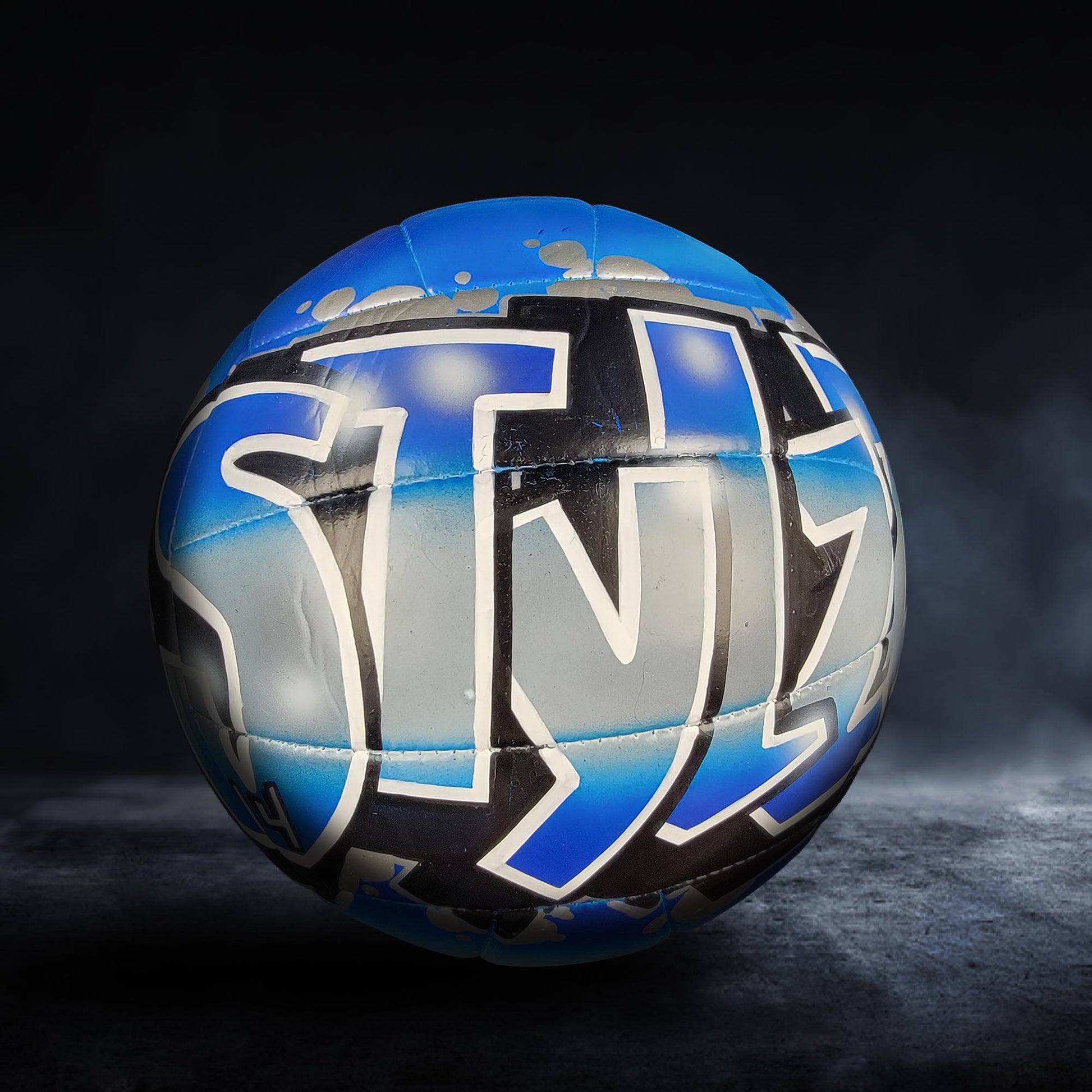 A blue and black sports ball with graffiti-style text on it, set against a dark background.