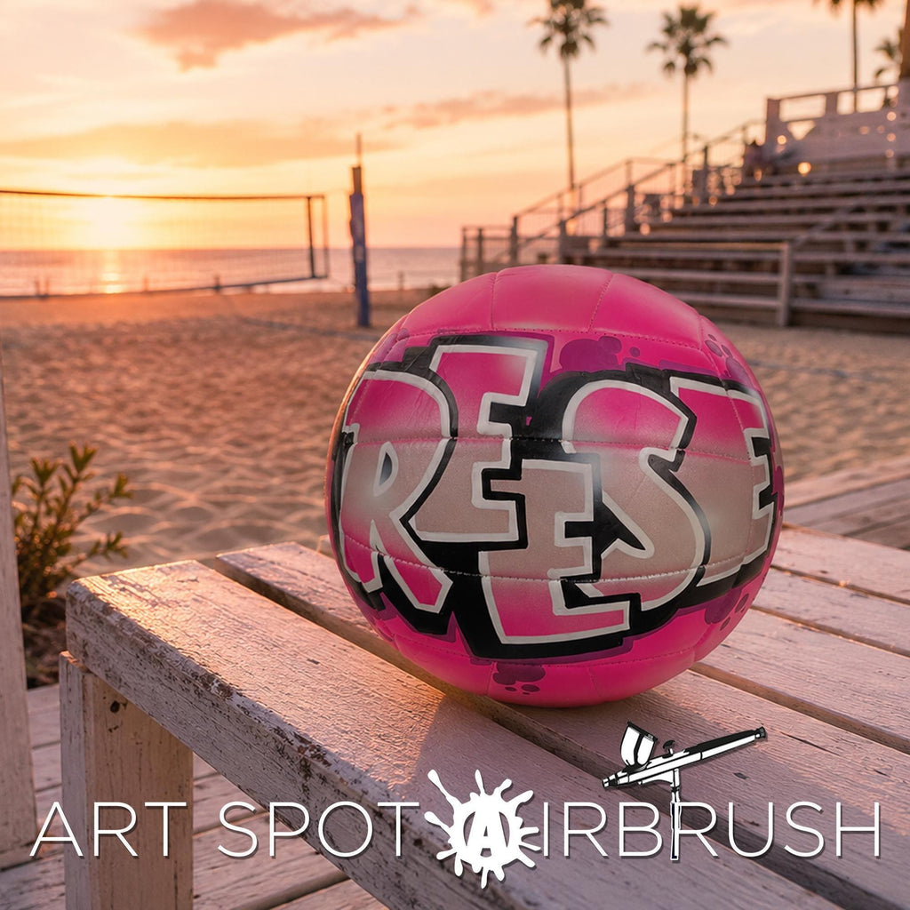 A pink custom volleyball personalized with the word "REESE" painted on it, sitting on a wooden bench near a beach at sunset.