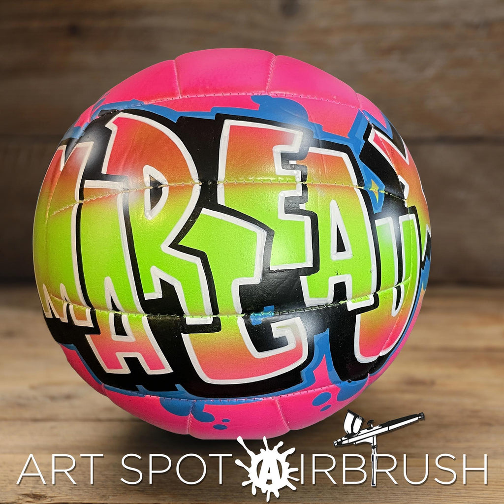 A colorful, graffiti-style personalized custom volleyball with the word "MARGEAUX" painted on it, sitting on a wooden surface.