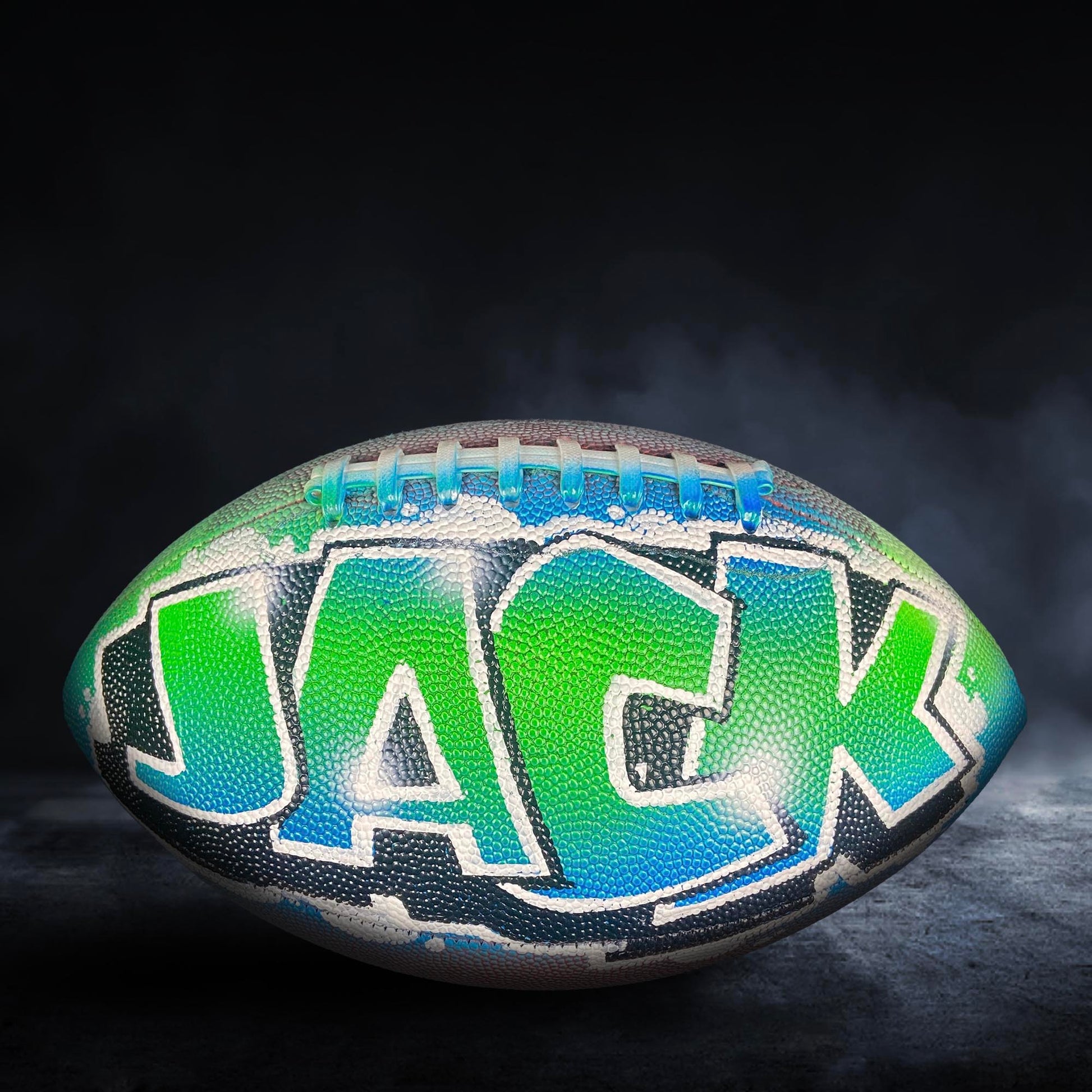 A colorful football with the word "JACK" painted on it, set against a dark background with smoke or fog.