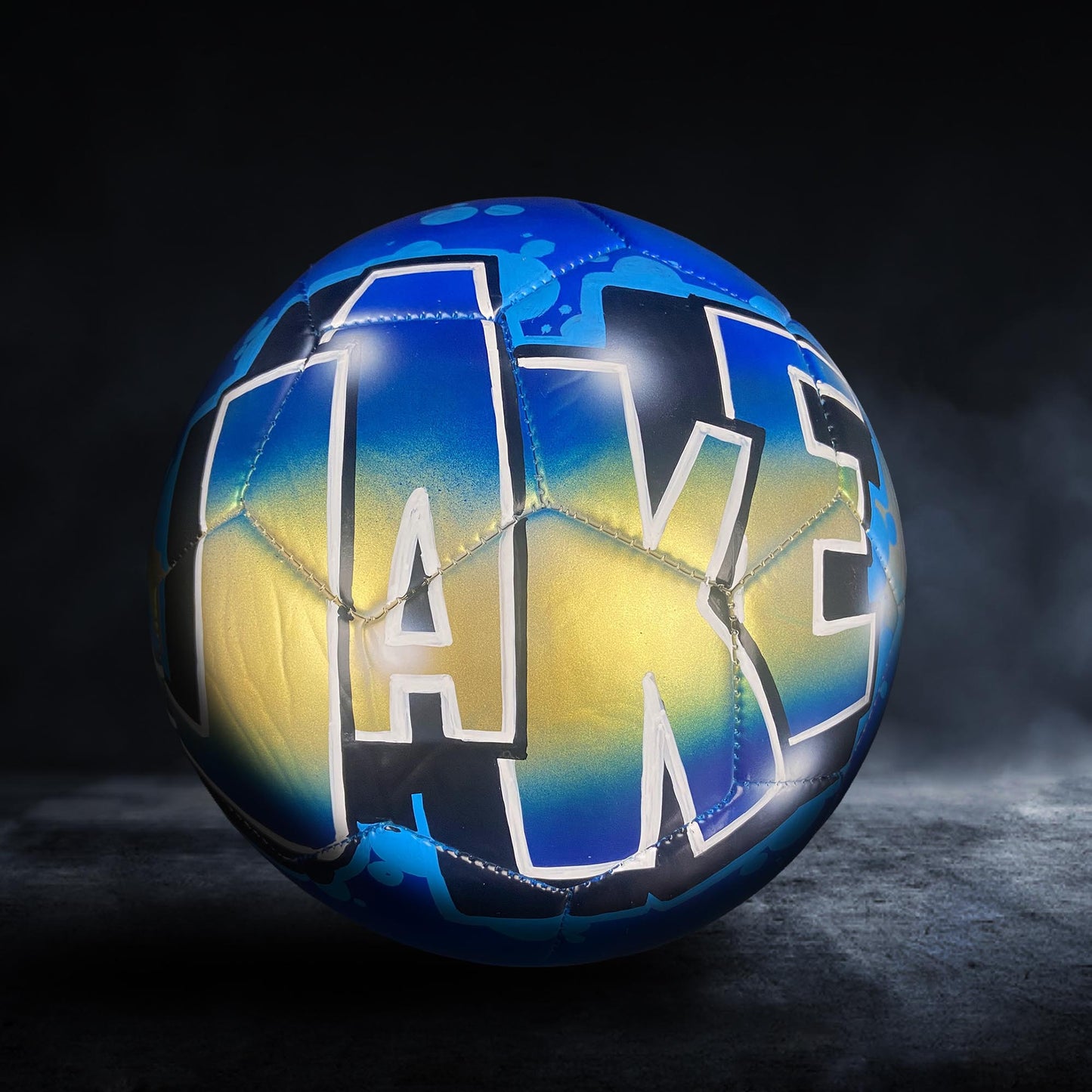 A soccer ball with the word "Lake" painted on it in blue and yellow colors.