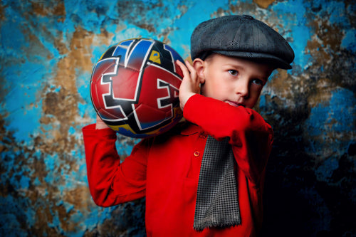 A young boy wearing a red shirt and a black cap is holding a colorful soccer ball in front of a blue and brown textured background.