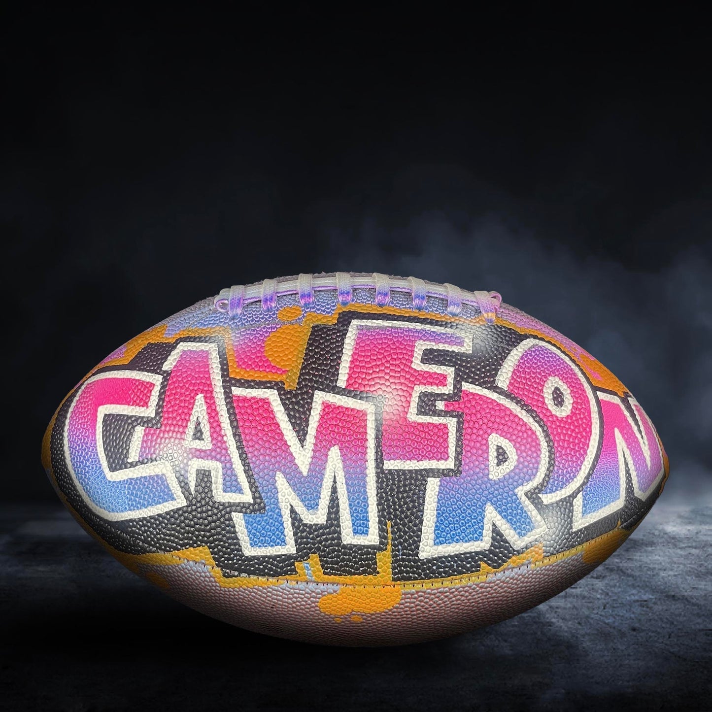 A colorful football with the name "CAMERON" painted on it, against a dark background with smoke or fog.