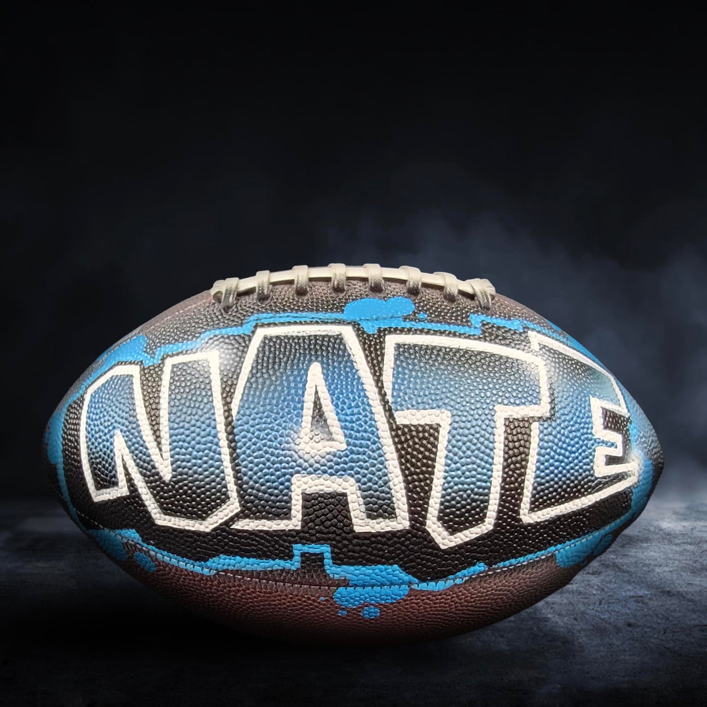 A football with the word "NATE" painted on it, against a dark background with smoke.