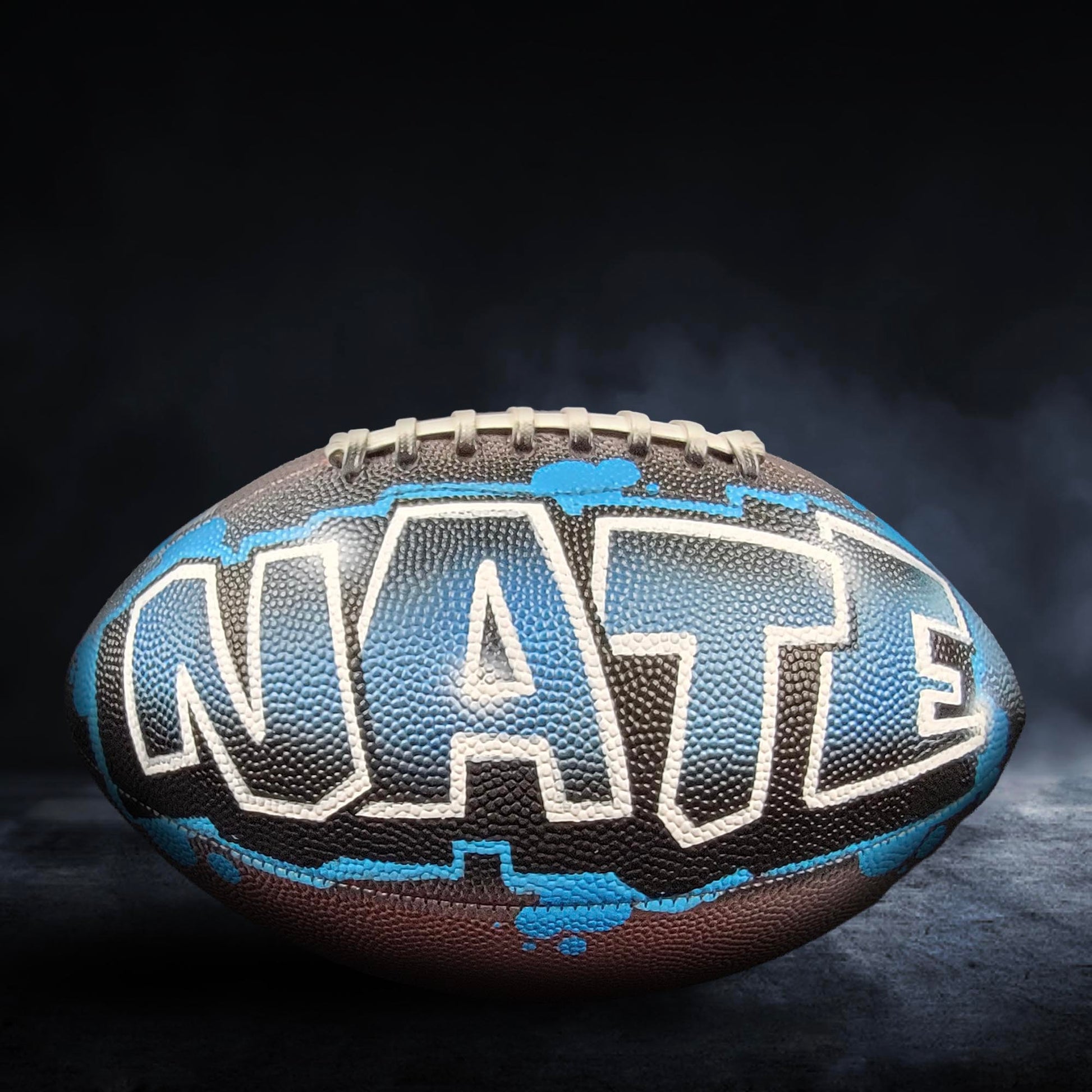 A football with the word "NATE" painted on it, against a dark background with smoke.