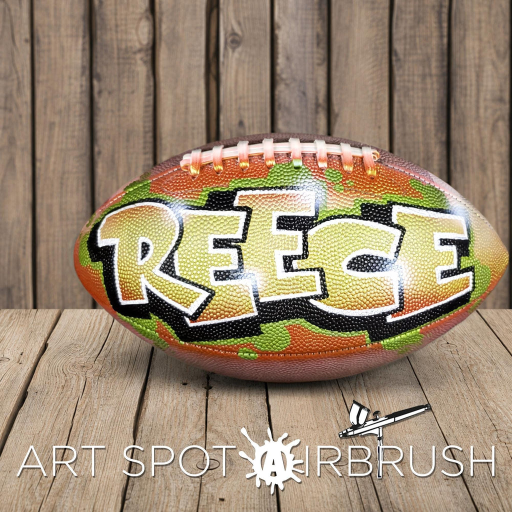 A football with the name "REECE" painted on it, sitting on a wooden surface.