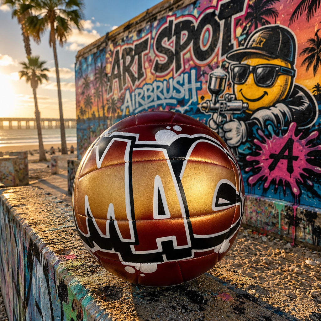 A red and gold custom volleyball personalized with the letters "MAC" on it, placed on a concrete ledge in front of a colorful ART SPOT AIRBRUSH graffiti mural.
