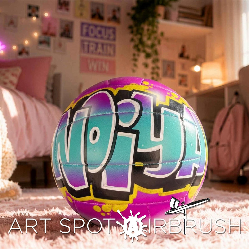 A colorful, graffiti-style personalized custom volleyball with the word "NOIYA" painted on it, sitting on a pink carpet in a bedroom with various decorations and furniture.
