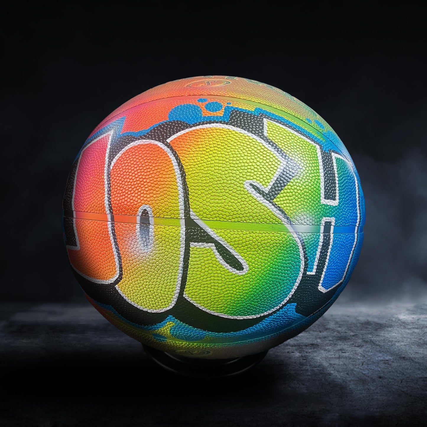 A colorful basketball with the name "Josh" painted on it, sitting on a dark surface.