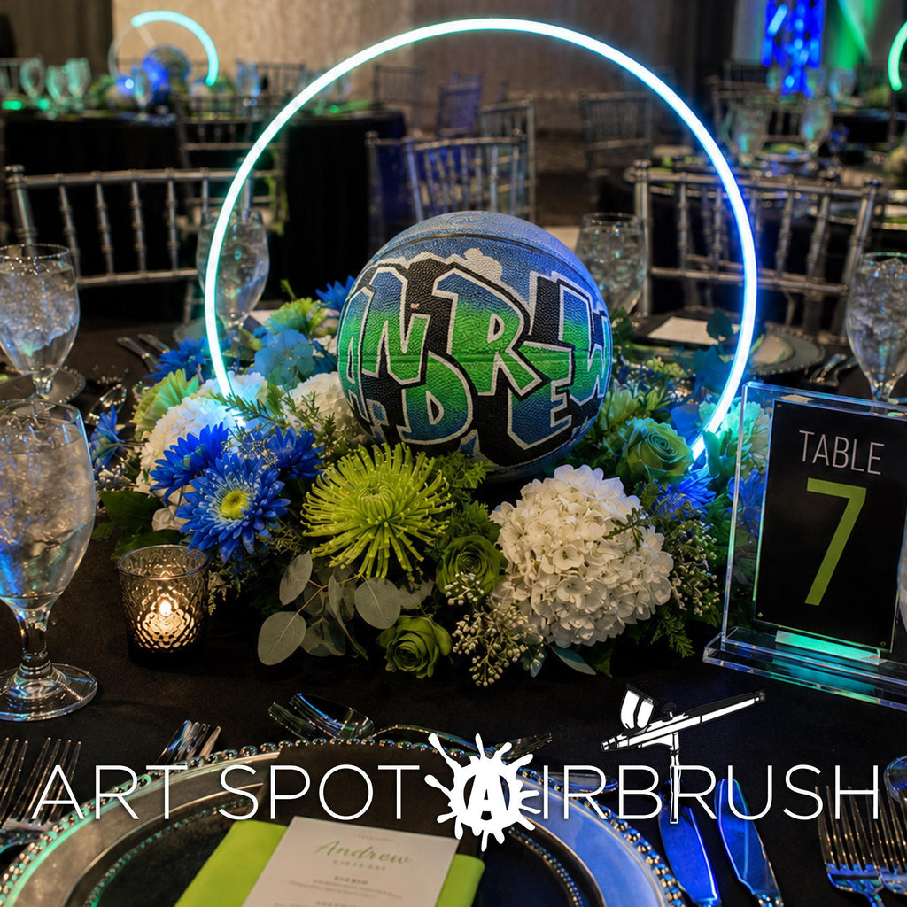 a table setting with a centerpiece of a basketball with graffiti on it, surrounded by flowers and greenery. The table is set with place settings, including wine glasses, forks, knives, and spoons. The table is also decorated with a circular light fixture and a table number sign.