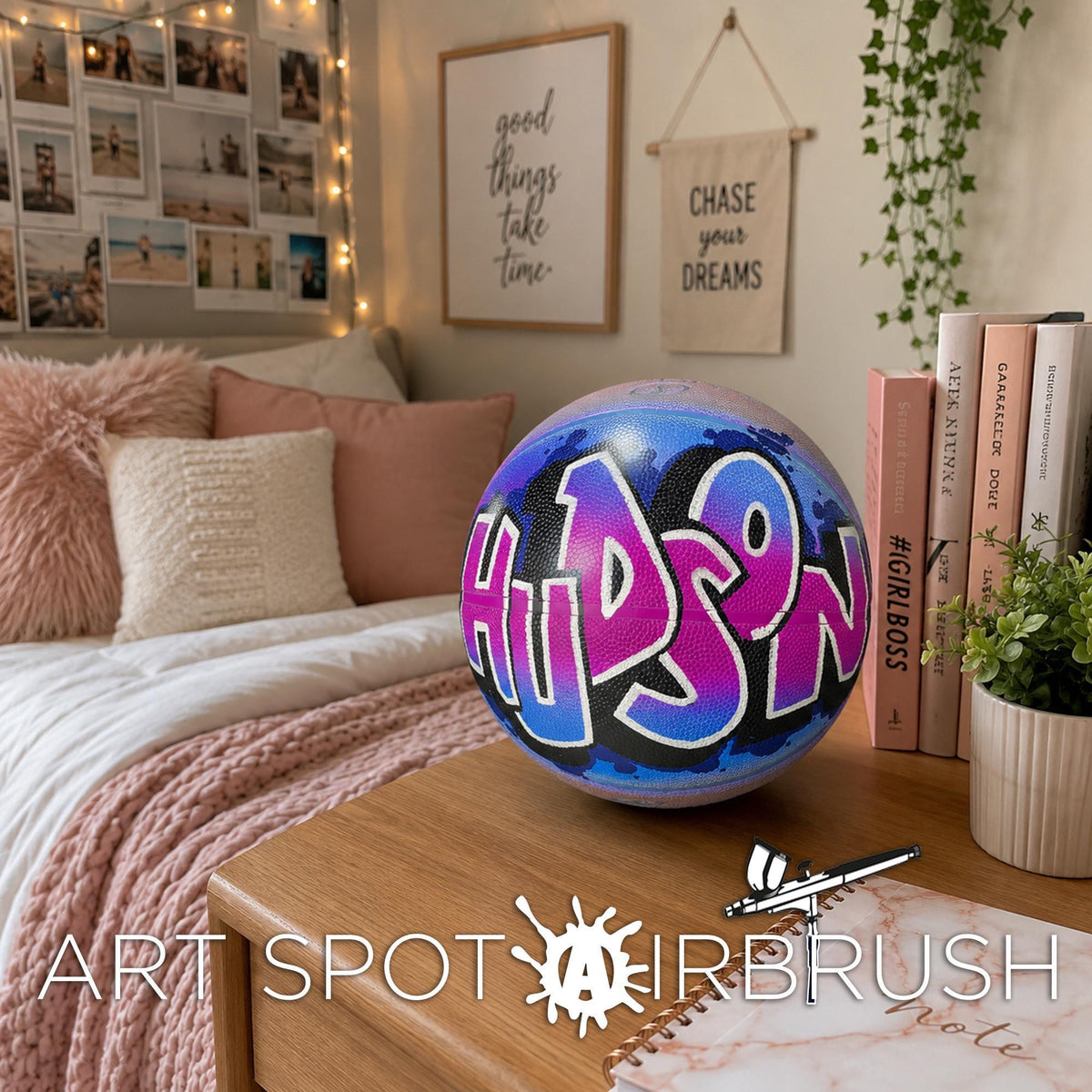 A bedroom with a bed, books, and a potted plant. The bed has a white comforter and a pink throw pillow. There is a basketball with the name "HUDSON" painted on it, and a sign that says "CHASE your DREAMS". The room also has a wall hanging with the phrase "good things take time" and a plant.