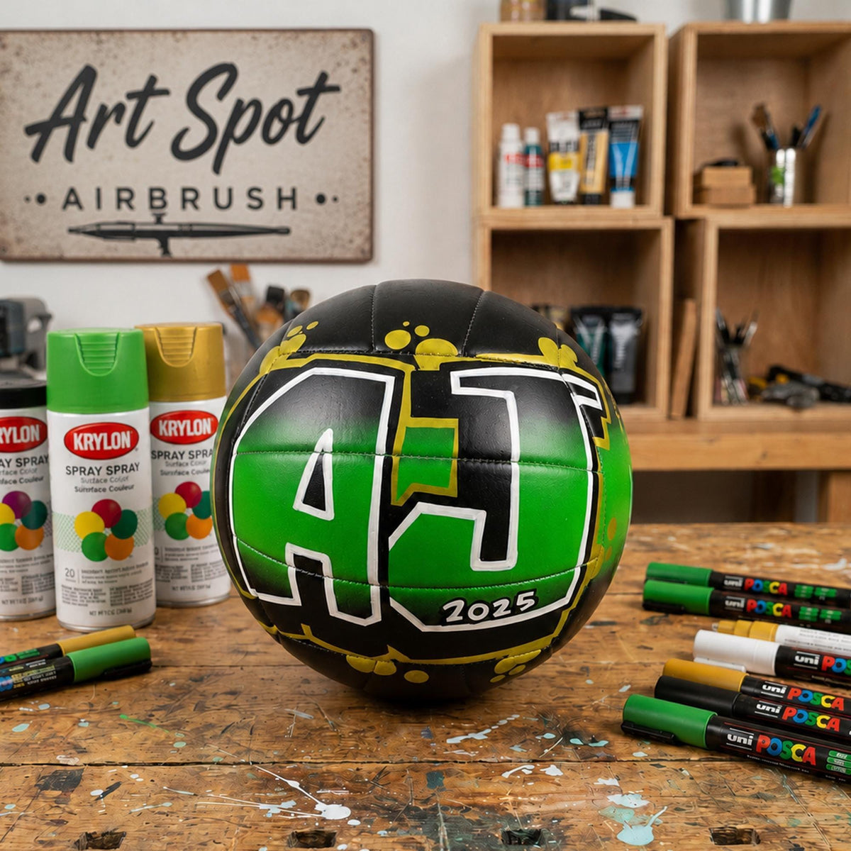 a black and green spray-painted custom volleyball personalized with the letters "AJ" on it, surrounded by various art supplies such as spray paint cans and markers on a wooden surface.