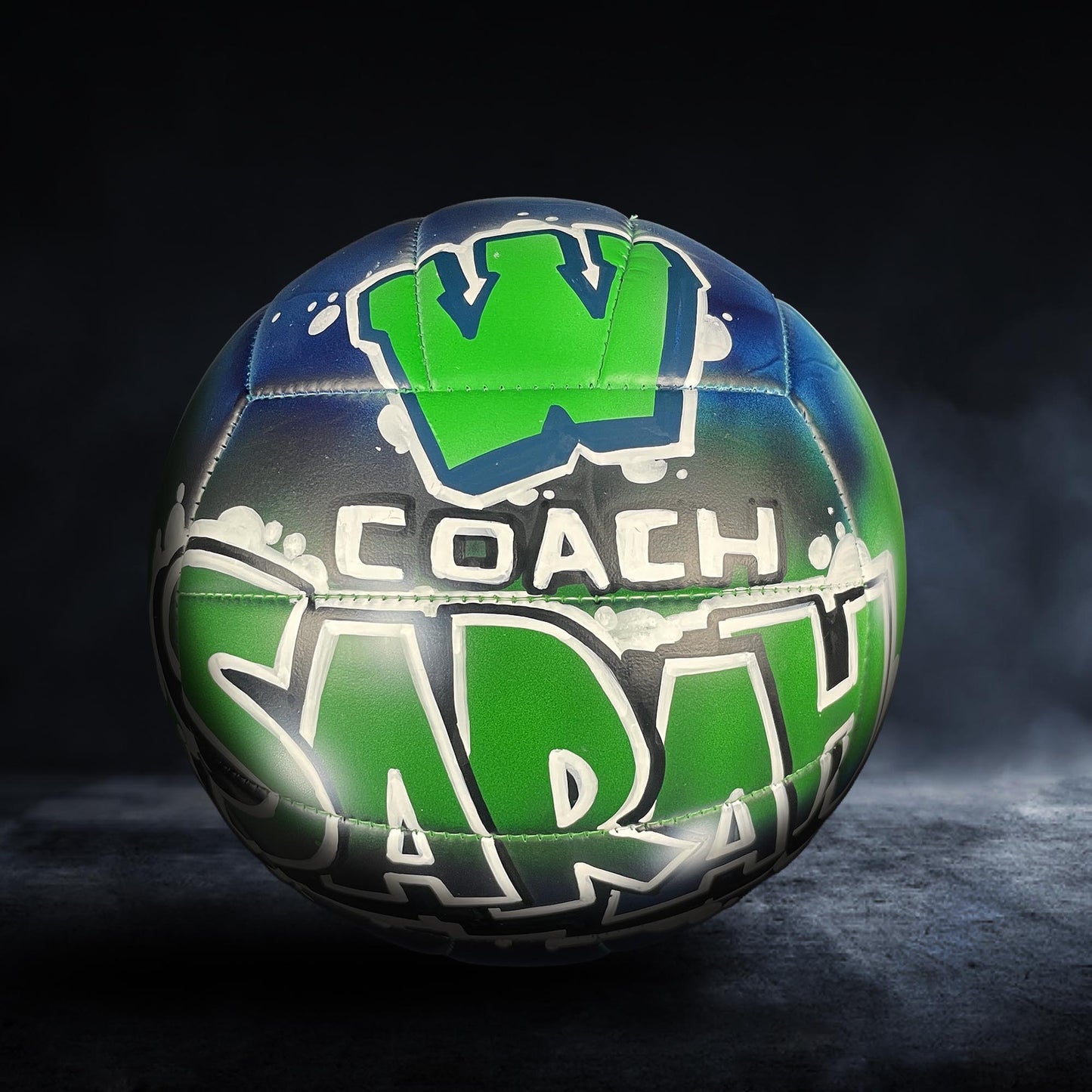 A colorful soccer ball with the word "COACH" painted on it, set against a dark background.