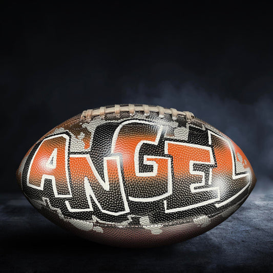 A football with the word "ANGEL" painted on it, against a dark background with smoke or fog.