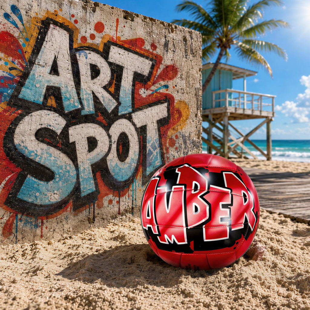 A red sports ball with the word "Amber" written on it, sitting on a sandy beach in front of a colorful graffiti wall.