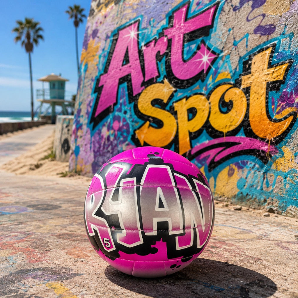 A pink and black custom volleyball personalized with the word "RYAN" on it, sitting on a concrete surface in front of a colorful graffiti Art Spot mural on a wall.