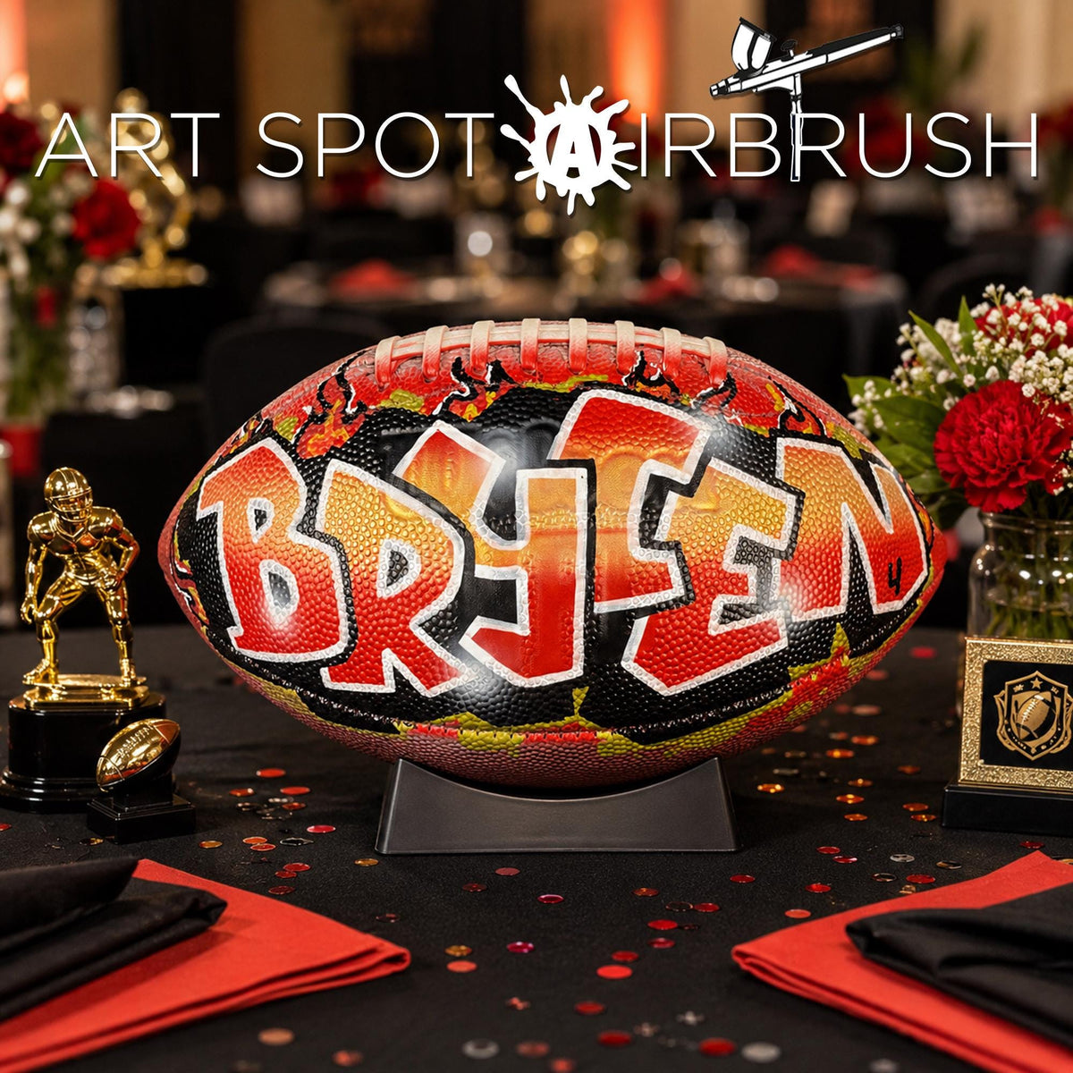a personalized custom football with the name "BRYCEN" painted on it, placed on a table with a trophy and flowers.