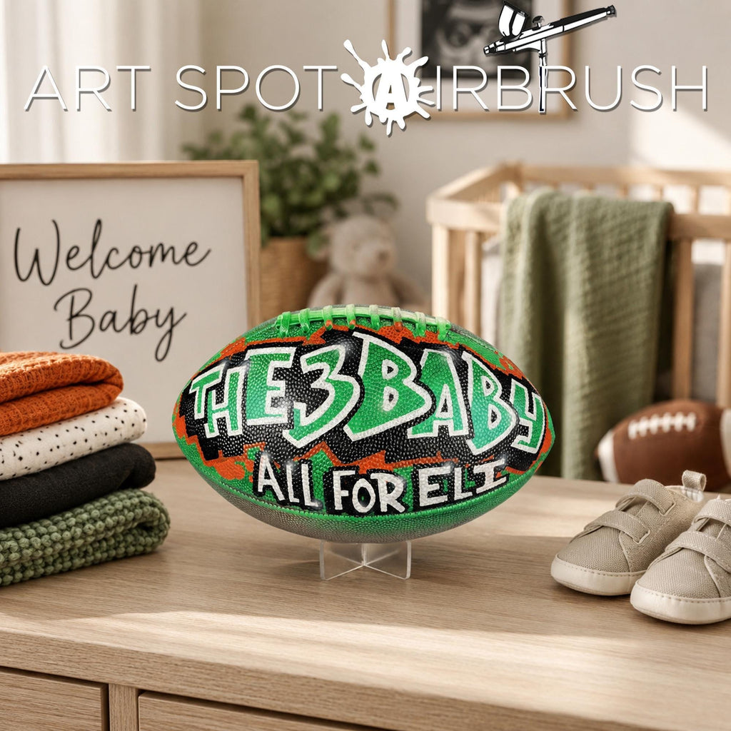a personalized custom football with the text "THE 3 BABY - ALL FOR ELI" painted on it, placed on a wooden surface next to baby clothes and baby shoes.