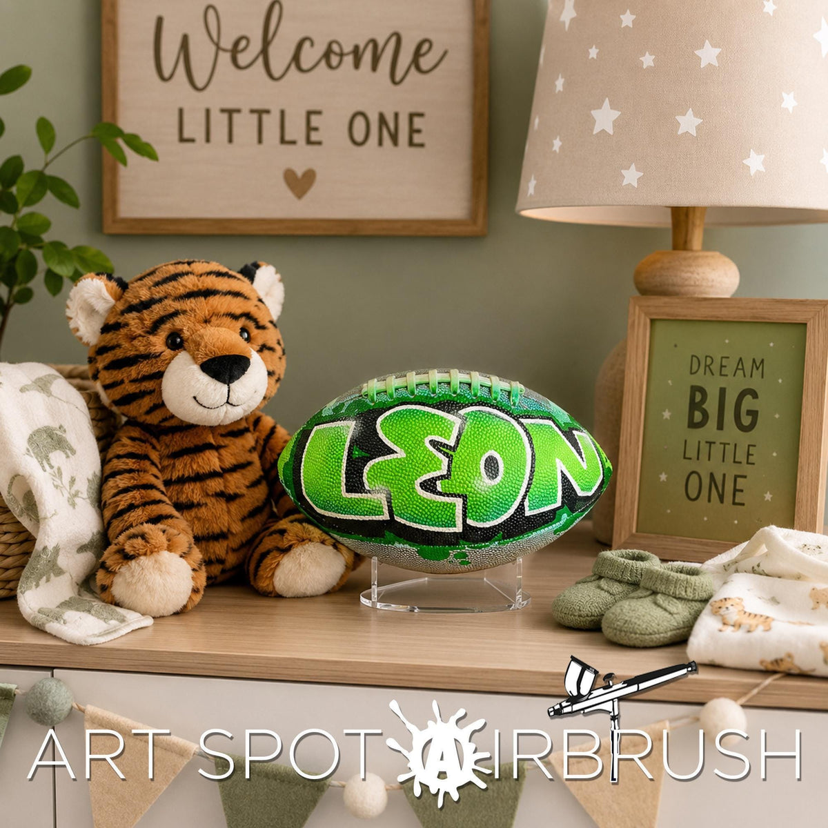 a stuffed tiger, a personalized custom football with the name "LEON" painted on it, and a framed sign that says "DREAM BIG LITTLE ONE". The objects are arranged on a wooden surface, with a lamp and a plant in the background.