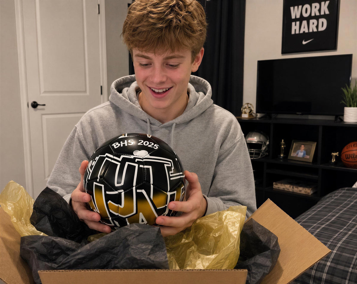 A young man is holding a soccer ball with the text "BHS 2023" printed on it, smiling as he looks at the ball.