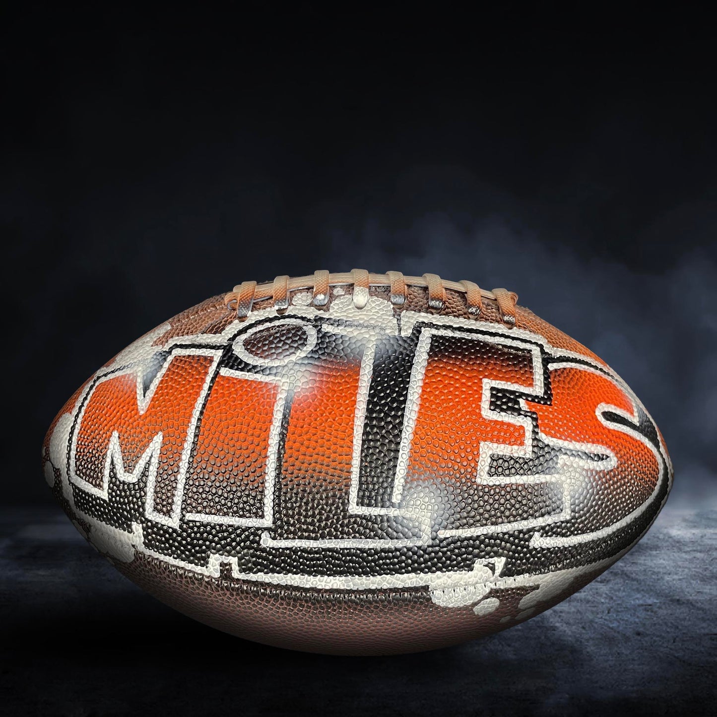 A football with the word "MITS" written on it, against a dark background with smoke or fog.
