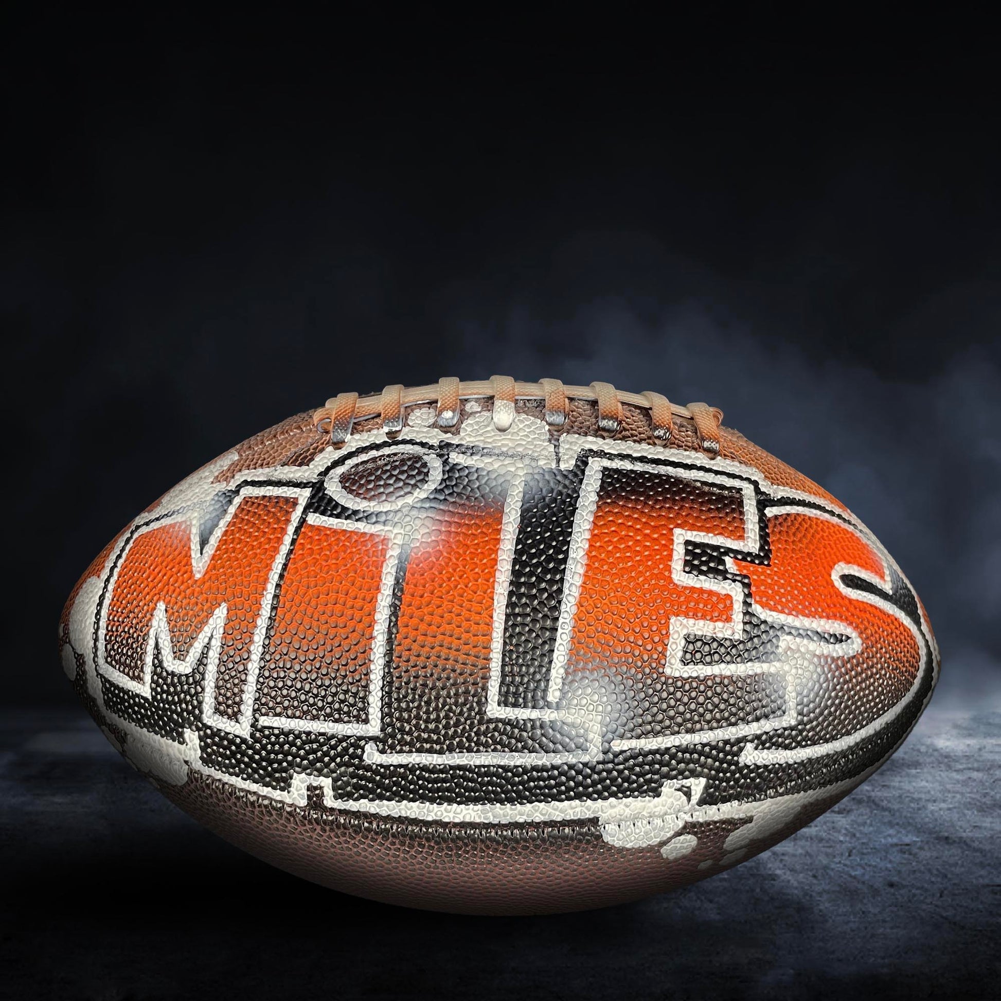 A football with the word "MITS" written on it, against a dark background with smoke or fog.