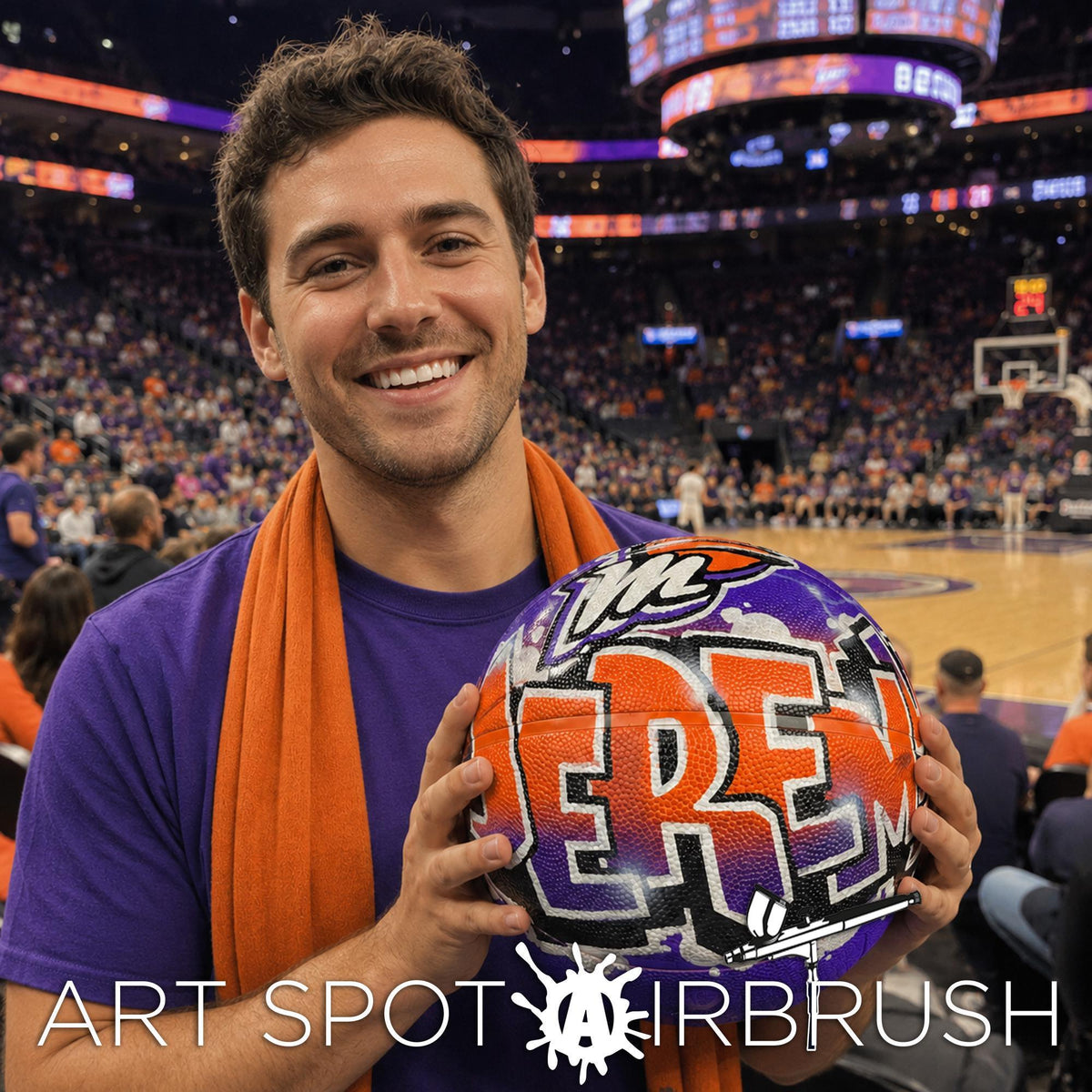 A man holding a basketball with the word "REB" on it, smiling and posing in front of a basketball court with a crowd in the background.